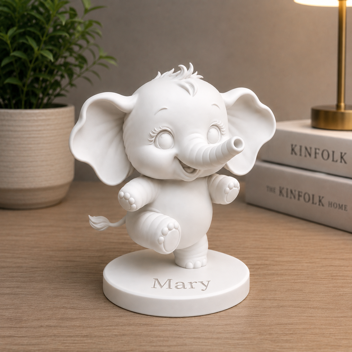 A white personalized elephant figurine standing on a round base engraved with the name 'Mary', placed on a wooden tabletop with a potted plant and books in the background.