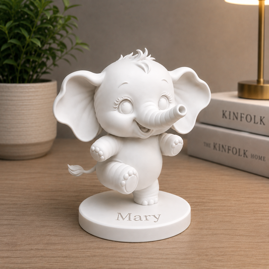 A white personalized elephant figurine standing on a round base engraved with the name 'Mary', placed on a wooden tabletop with a potted plant and books in the background.