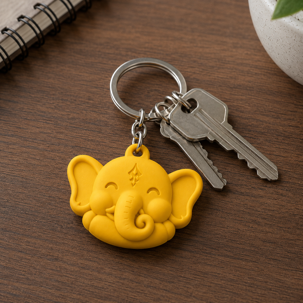 A yellow elephant-shaped keychain with attached silver metal keys, photographed on a wooden tabletop with a notebook and small plant visible in the background.