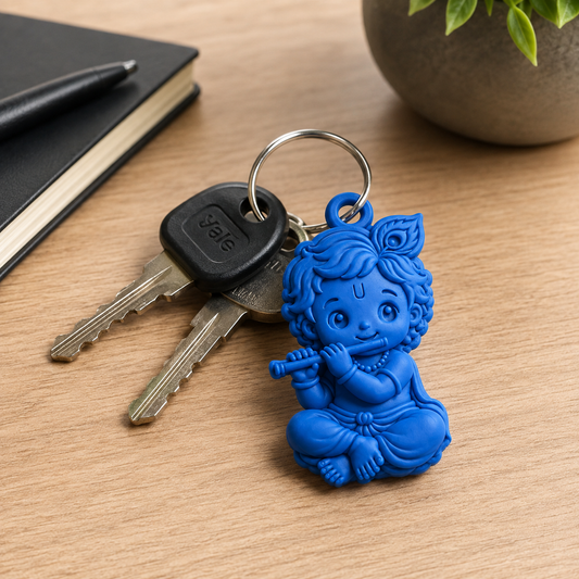 A blue plastic keychain shaped like a seated child playing a flute, attached to a metal keyring with two keys. The keychain is photographed on a wooden tabletop with a notebook, pen, and plant visible in the background.