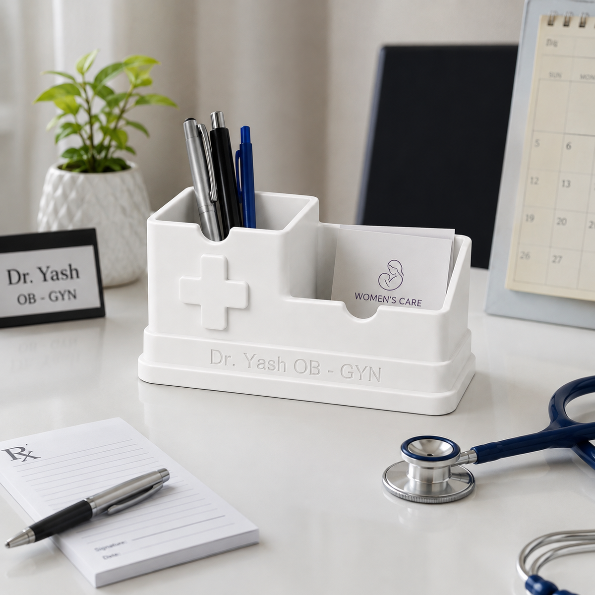 A white plastic dual-compartment desk organizer with a medical cross emblem and the text 'Dr. Yash OB - GYN' on the front, holding pens and a business card, styled on a doctor's office desk with a stethoscope, notepad, and plant.
