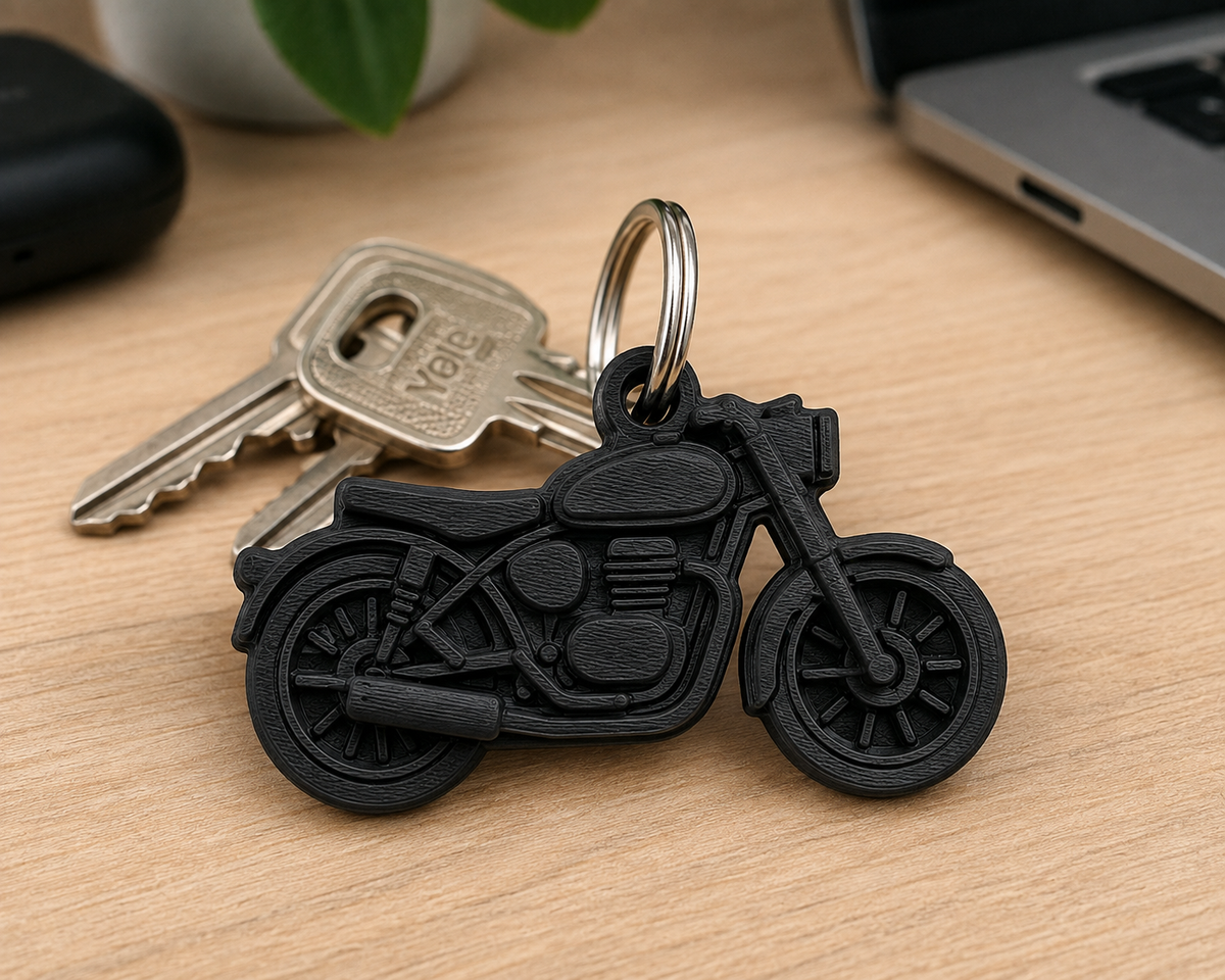 A black motorcycle-shaped keychain with attached metal keys, photographed on a light wood desk with blurred laptop, plant, and case in the background.