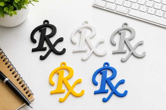 Five RX letter-shaped charms in black, white, gray, yellow, and blue, arranged on a white desk with a keyboard, notebook, pen, and small plant visible.